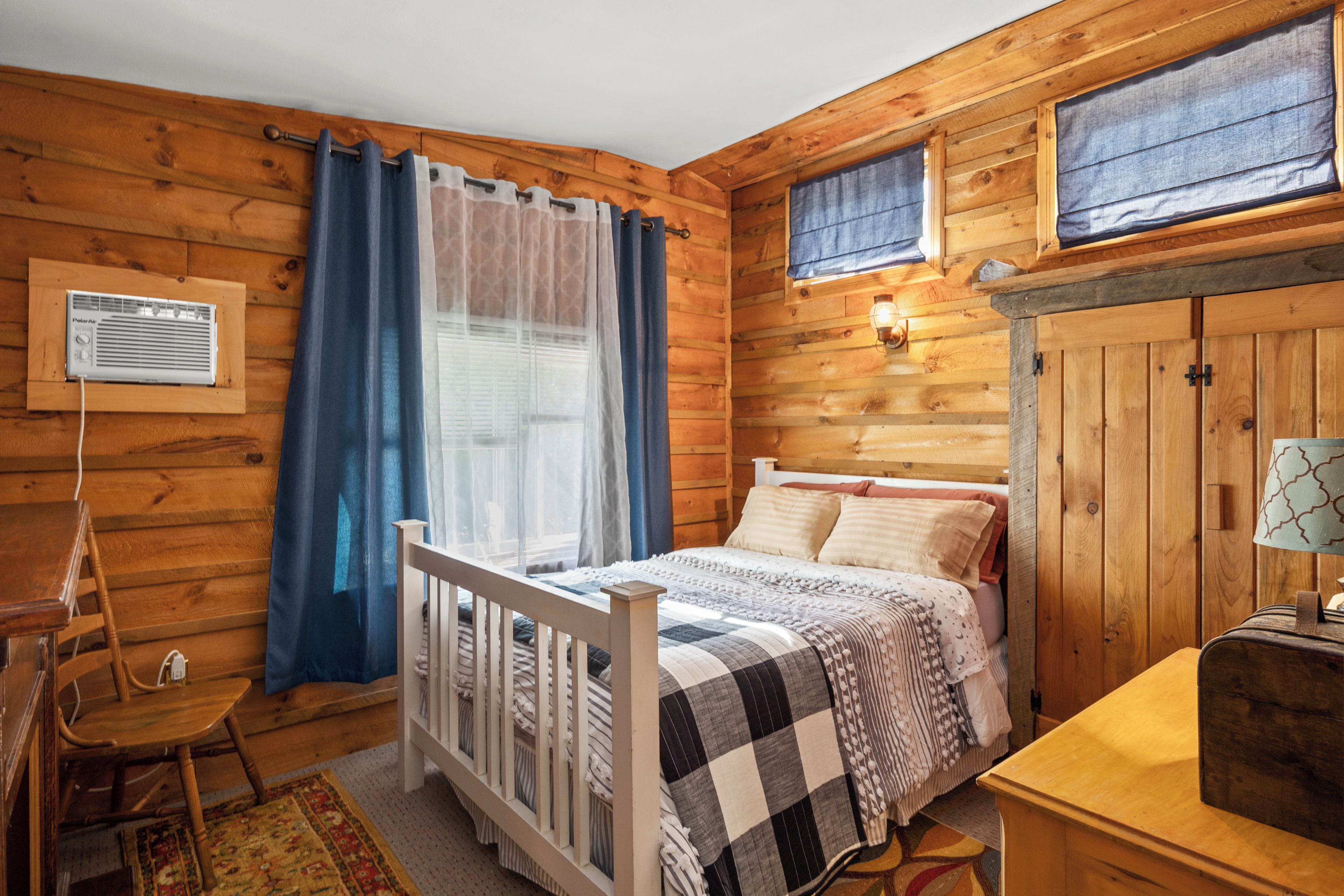 Dormitorio de cabaña de madera con cama, escritorio, silla, ventana con cortinas y calentador montado en la pared.