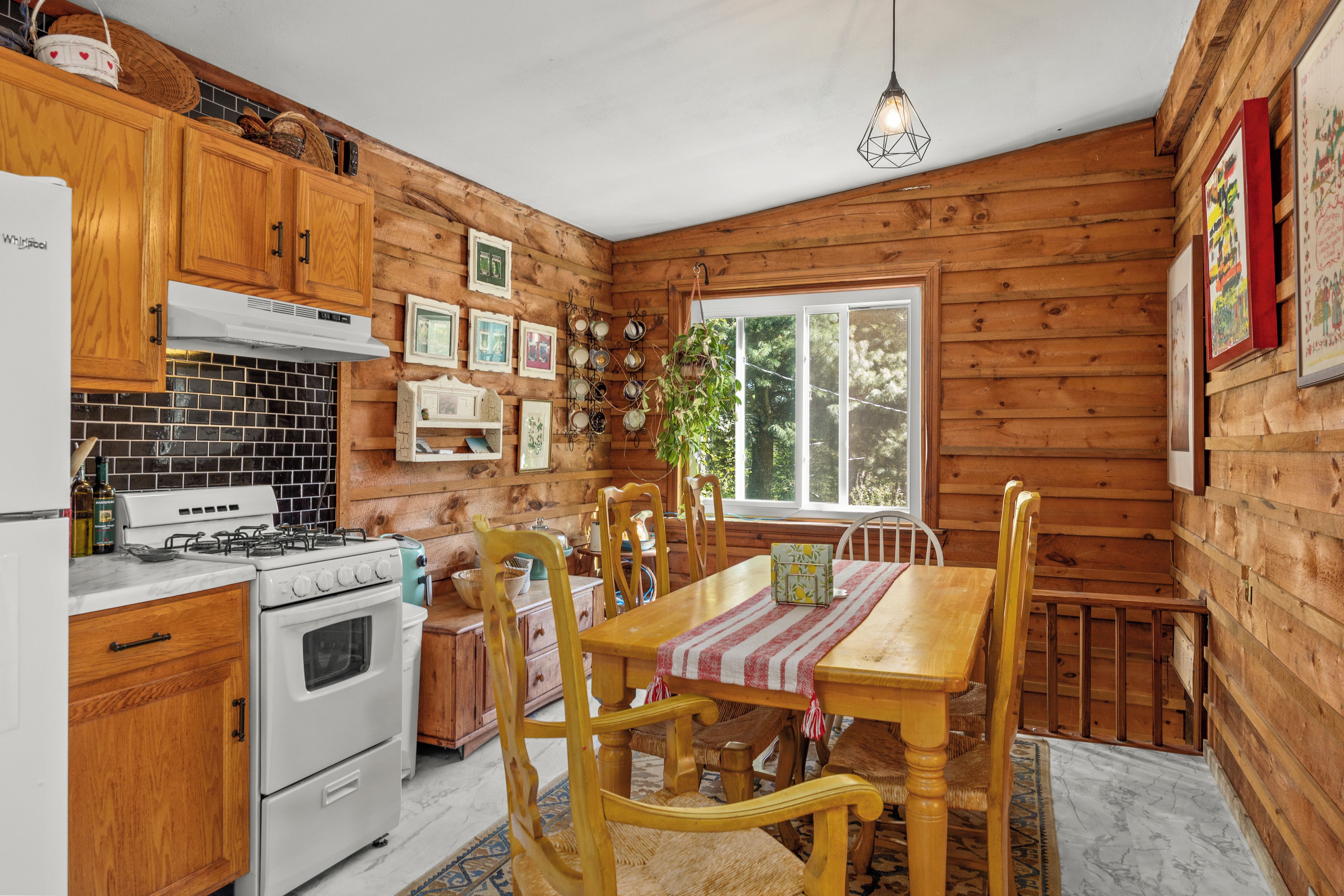 A wooden cabin interior with a kitchen, dining table, chairs, and a hanging light fixture.