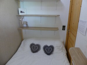 A bed with two heart-shaped pillows, a wooden shelf above, a small chair, and a door with a paper on the wall. - Studio-cabin near slopes - 4 guests (Pralognan-la-Vanoise)
