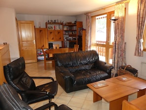 Een woonkamer met een leren bank, een houten bijzettafeltje en bijpassende stoelen. Een boekenplank is gevuld met boeken en decoratieve objecten.