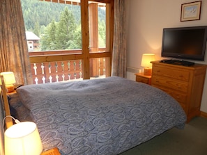 A bedroom with a bed, a television on a wooden dresser, and a window with a view of trees. - Comfortable apartment - 7 guests (Pralognan-la-Vanoise)