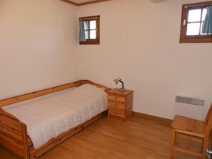 A single bed with a wooden frame and a white bedspread, a small wooden nightstand with a lamp, a radiator, and a window with a curtain. - Cozy three-room near slopes - 6 guests (Pralognan-la-Vanoise)