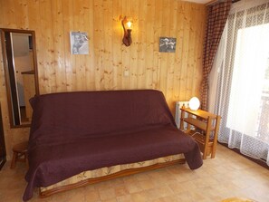 A room with a wooden wall, a bed with a purple bedspread, a small table with a lamp, and a window with curtains. - Convenient studio near slopes - 4 guests (Pralognan-la-Vanoise)