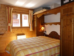 A wooden room with a bed, a window with curtains, a lamp, and a wooden headboard. - Cozy mountain-style apartment - 4 guests (Pralognan-la-Vanoise)