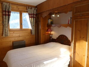 Una habitació de fusta amb un llit, una finestra amb cortines, una làmpada i una estant amb llibres.