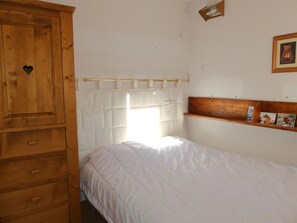 Ein Schlafzimmer mit Holzschrank, einem Bett mit weißer Bettwäsche, einem Fenster mit weißem Vorhang und einer Holzablage mit Bildern.