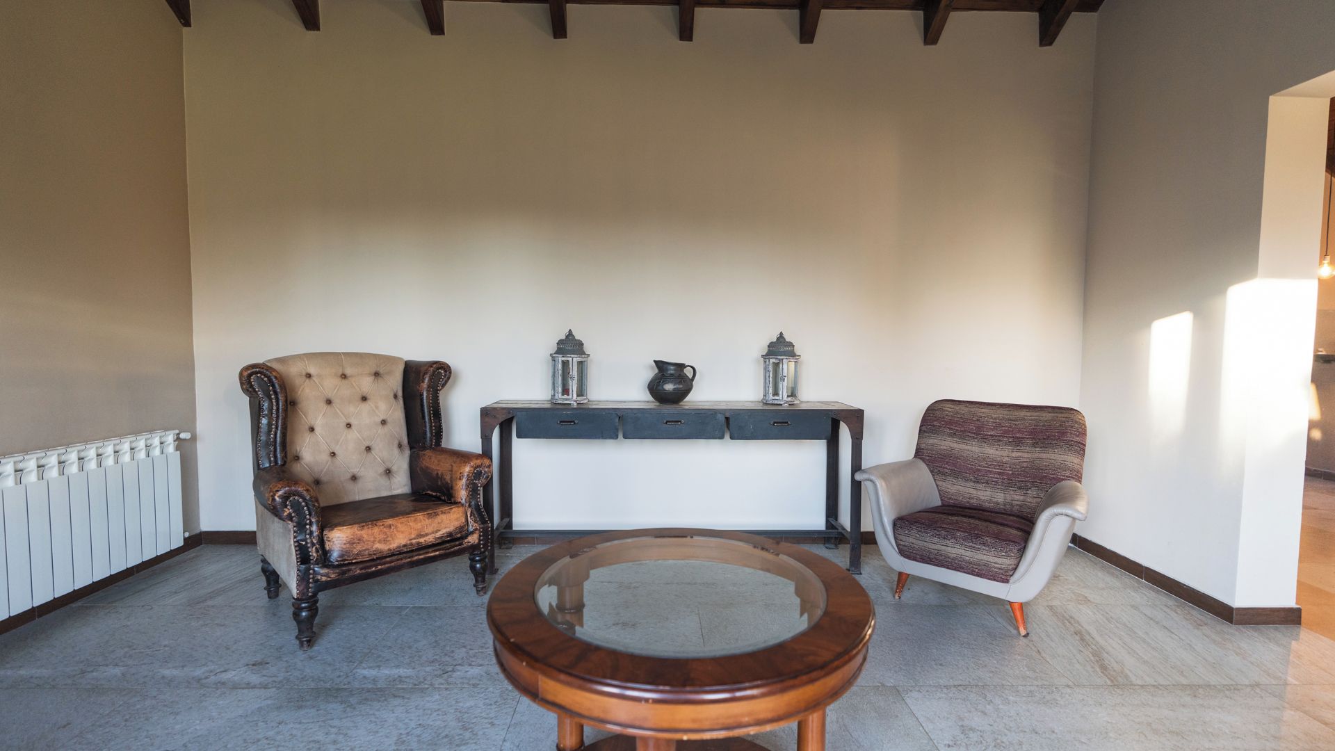 A spacious room with a high ceiling, a round wooden table, two upholstered chairs, a small console table with decorative items, and a radiator along the wall.