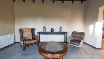 A spacious room with a high ceiling, a round wooden table, two upholstered chairs, a small console table with decorative items, and a radiator along the wall.