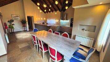 A dining area with a long table, red and blue chairs, a kitchen in the background, and a ceiling with exposed beams and hanging lights.