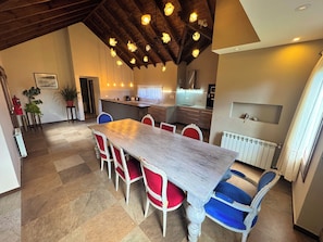 Uma área de refeições com uma mesa comprida, cadeiras vermelhas e azuis, uma cozinha ao fundo e um teto com vigas aparentes e luminárias pendentes.