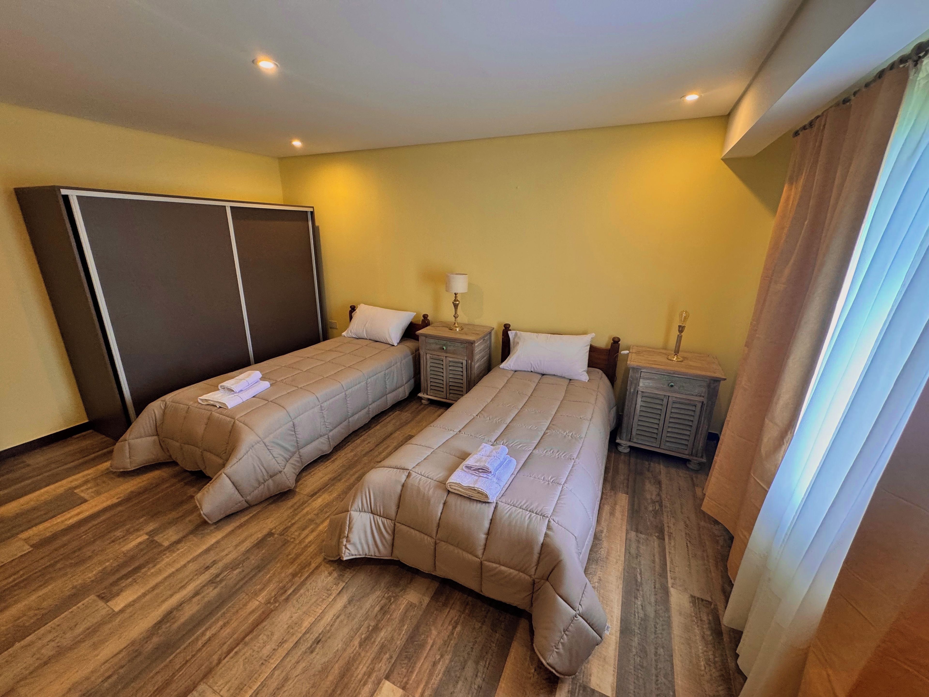 Habitación de hotel con dos camas, piso de madera, un armario, mesitas de noche con lámparas y una ventana con cortinas.