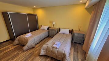 Habitación de hotel con dos camas, piso de madera, un armario, mesitas de noche con lámparas y una ventana con cortinas.