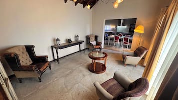 A living room with a sofa, armchair, and a round wooden table. There is a bench with decorative items and a view into a dining area with chairs and a table.