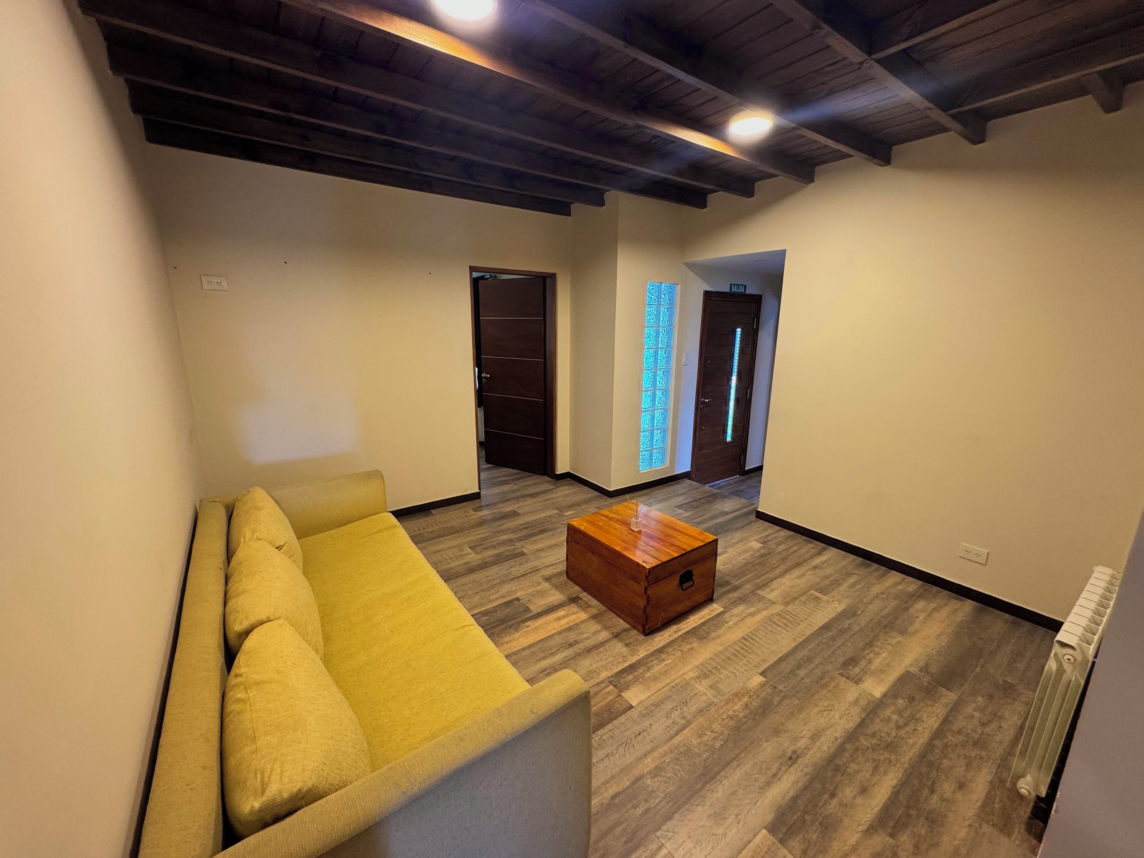 Una sala de estar moderna con un sofá amarillo, una mesa de centro de madera y piso de madera. Se ven dos puertas, una que da a un pasillo y otra a una habitación con un elemento de iluminación azul.
