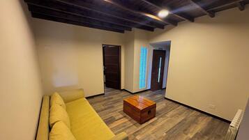 Una sala de estar moderna con un sofá amarillo, una mesa de centro de madera y piso de madera. Se ven dos puertas, una que da a un pasillo y otra a una habitación con un elemento de iluminación azul.
