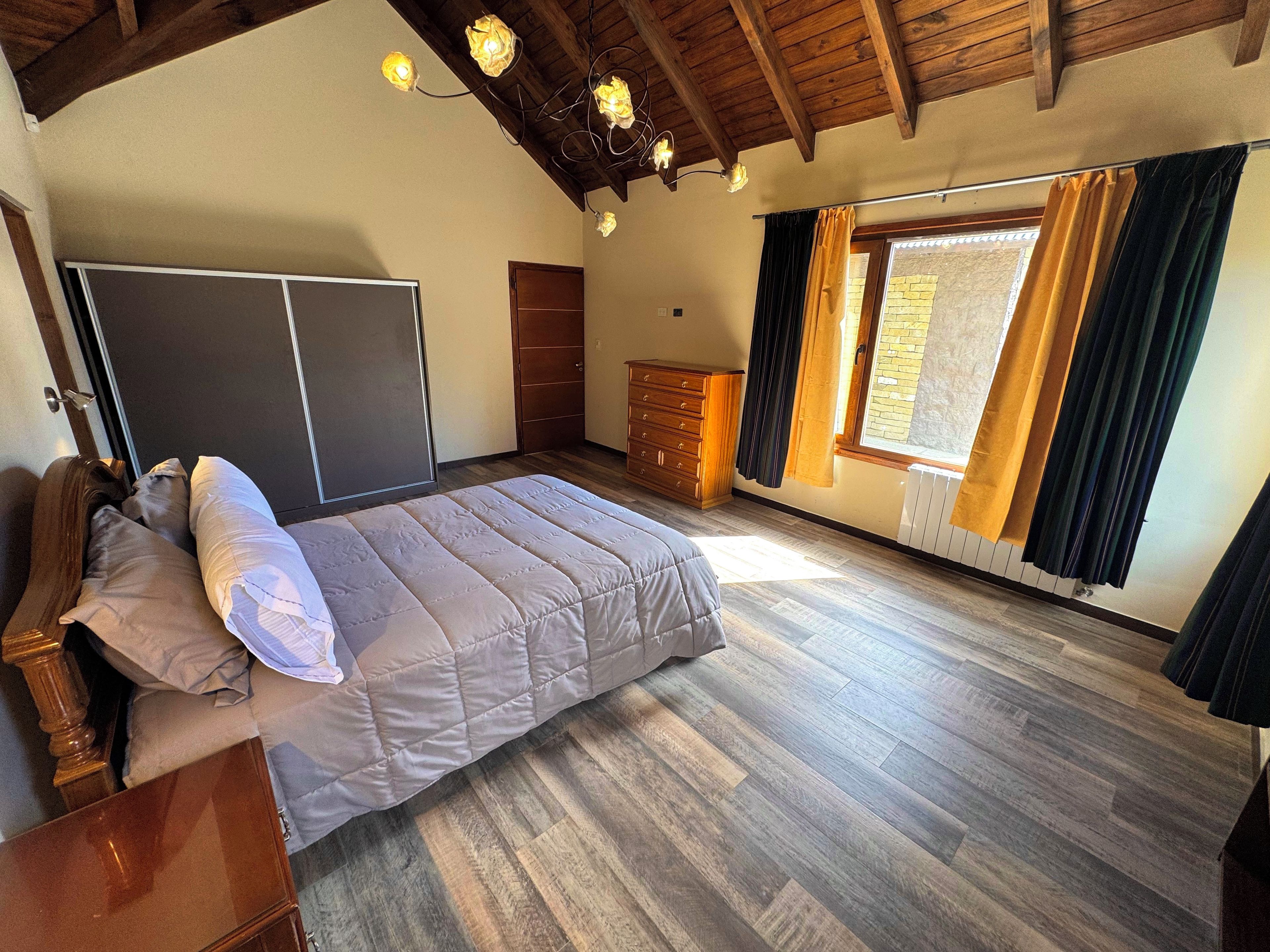 A bedroom with a bed, wooden headboard, a dresser, a wardrobe, and a window with curtains.