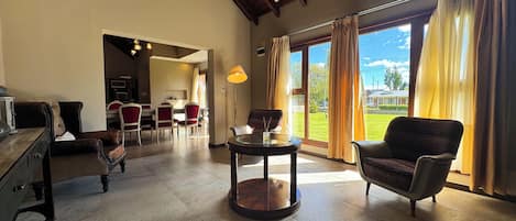 A spacious living area with a high ceiling, a dining area with red chairs, and a view of a green landscape through large windows.