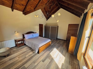 Quarto com teto de madeira, uma cama, mesa de cabeceira, guarda-roupa e janela com cortinas.