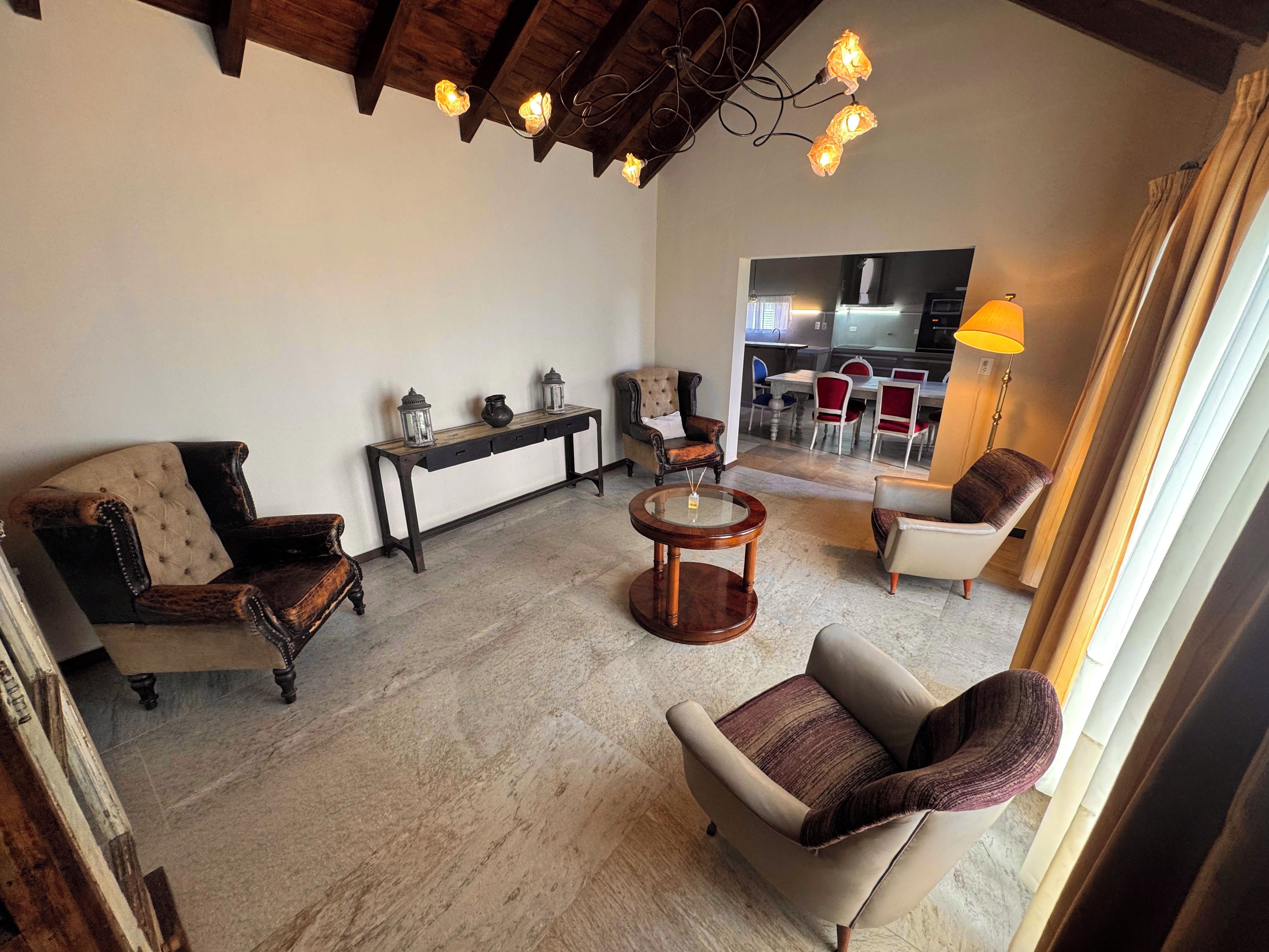 A living room with a sofa, armchair, and a round wooden table. There is a bench with decorative items and a view into a dining area with chairs and a table.