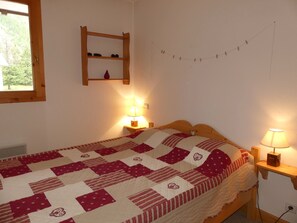 A bedroom with a wooden bed, a quilt, two bedside lamps, a shelf with a red object, and a window with a view of trees. - Ideal apartment at the slope base - 4 guests (Pralognan-la-Vanoise)