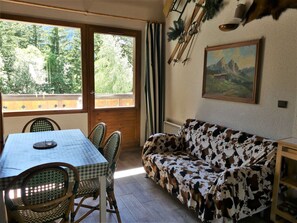 Ein Zimmer mit Tisch, Stühlen, einem Couchgarnitur mit Kuhmuster, einem Gemälde und einem Fenster mit Blick auf Bäume.