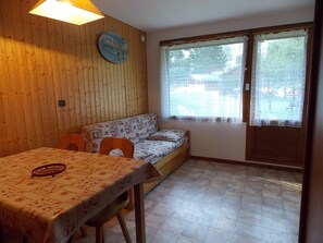 Een kamer met een houten wand, een bank, een eettafel met stoelen en een raam met gordijnen.