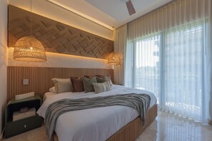 Quarto moderno com uma cama grande, duas luminárias de cabeceira, mesa de cabeceira e vista para o exterior através de cortinas translúcidas.