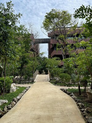 A paved pathway leading to a building with a modern architectural style, surrounded by lush greenery and a stone wall. - Lux 1BR  Ocean-View Pool  w/Habitas Beach Club (Tulum)