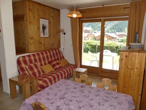 Ein gemütliches Zimmer mit einem karoüberzogenen Sofa, einem kleinen Holztisch, einem Bett mit einem blumigen violetten Bettbezug und Blick auf Häuser durch ein großes Fenster.