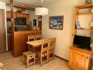 A compact kitchen with wooden cabinets, a dining table with chairs, a TV on a wooden stand, and a mountain painting on the wall. - Charming apartment without equipment - 4 guests (Pralognan-la-Vanoise)