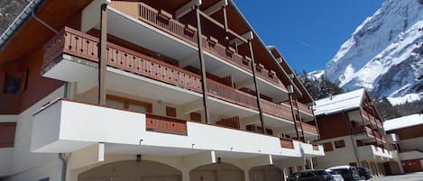 A multi-story hotel with balconies, a ski slope in the background, and a parking area.