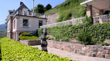 Een tweelaags huis met een balkon, omgeven door weelderige begroeiing en een stenen steunmuur.