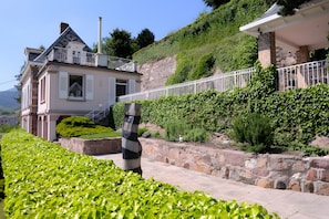 A two-story house with a balcony, surrounded by lush greenery and a stone retaining wall.