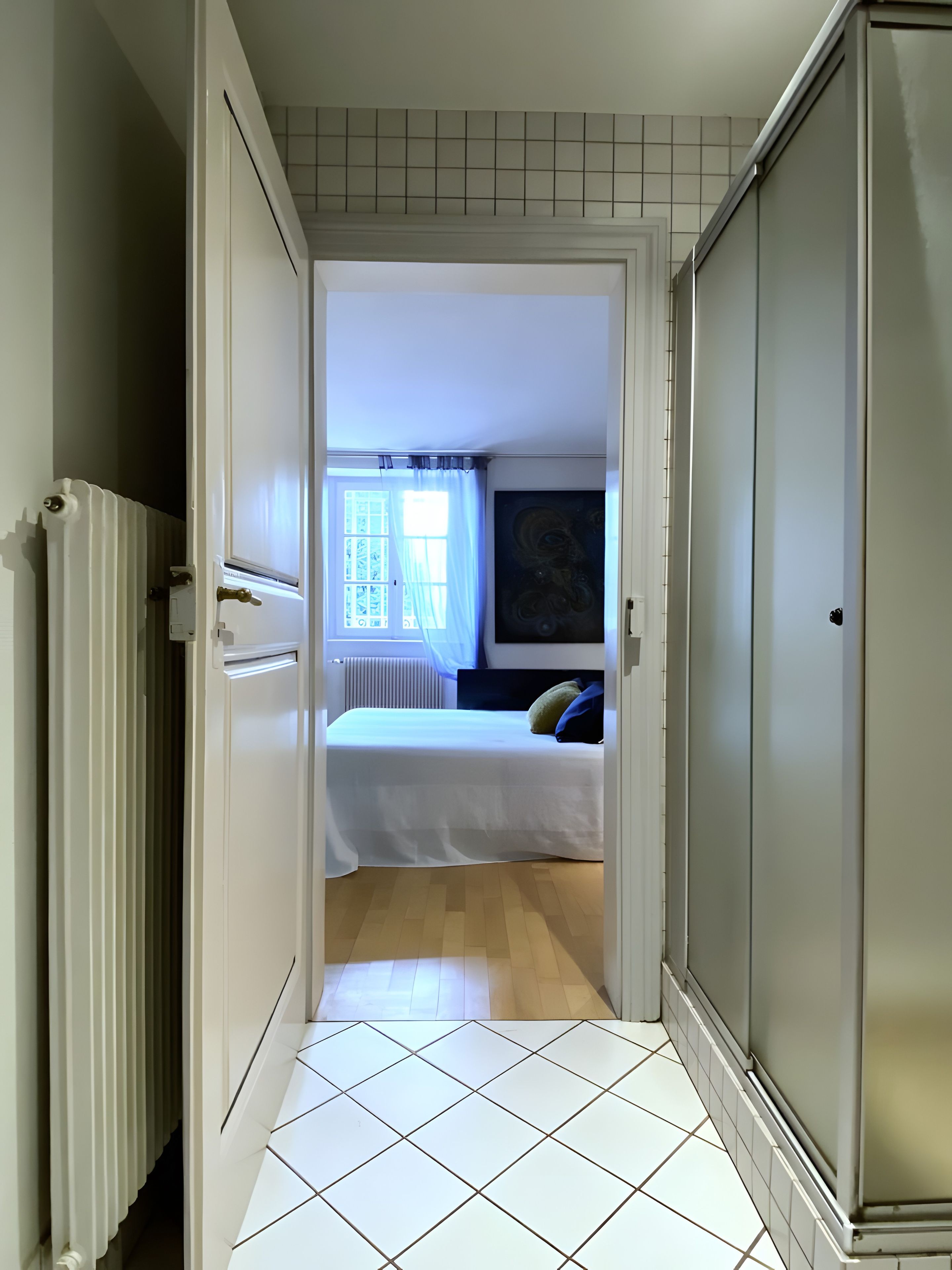 Een smalle gang die uitkomt in een slaapkamer met een bed, een radiator en een raam met gordijnen.