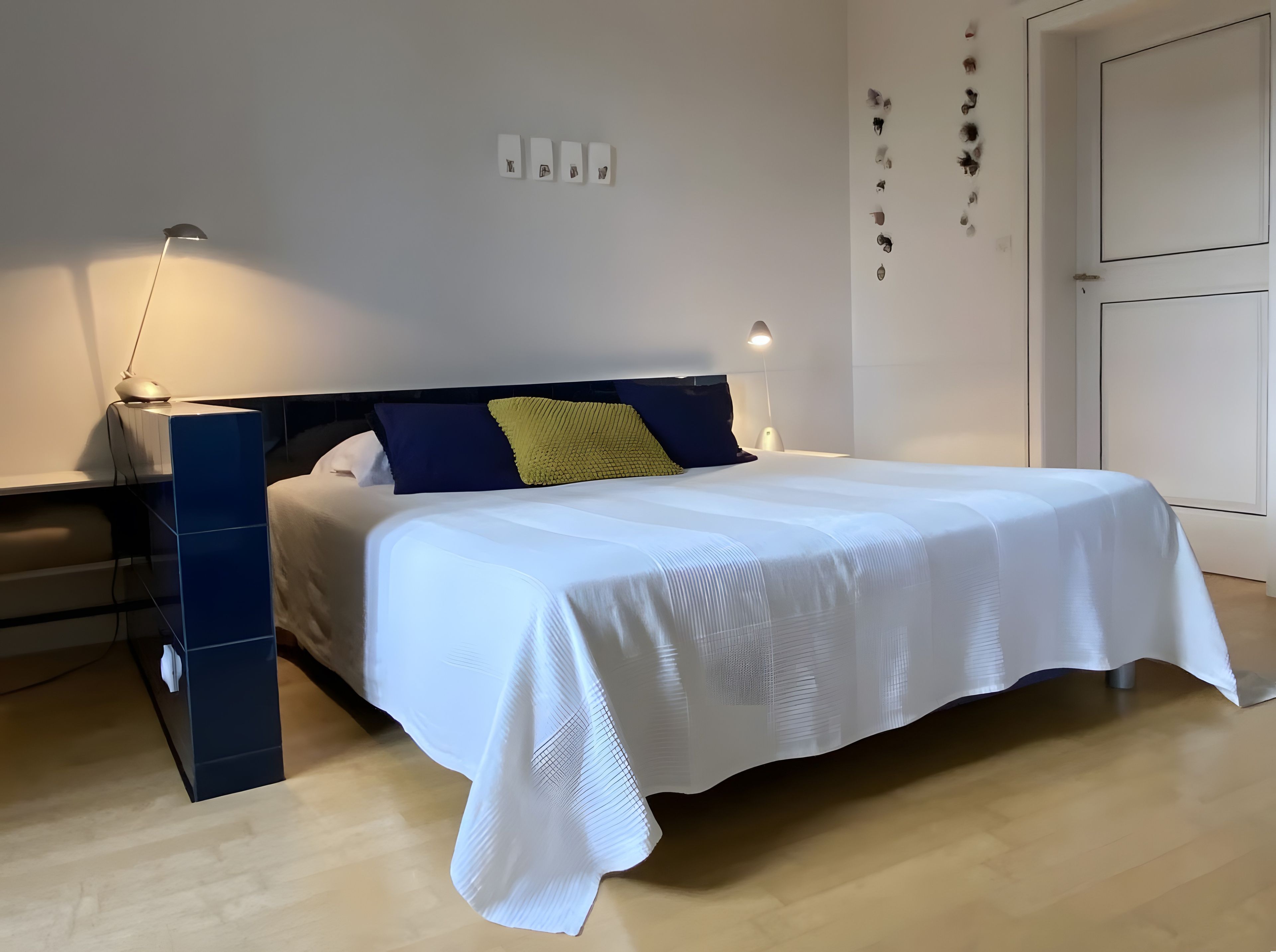 Een keurig opgemaakt bed met een witte dekbedovertrek, een nachtkastje met een lamp en een blauwe opbergkast.
