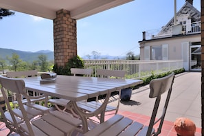 Een terras met een witte tafel en stoelen, een bakstenen pilaar en uitzicht op een huis met een witte hek en bergen op de achtergrond.