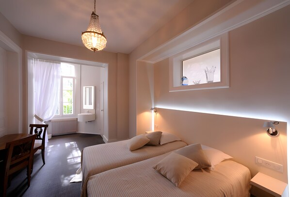 Een hotelkamer met twee bedden, een bureau, een stoel, een kroonluchter en een raam met uitzicht.
