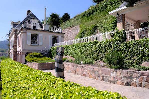 A two-story house with a balcony, surrounded by lush greenery and a stone retaining wall.