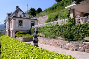 Una casa de dos pisos con balcón, rodeada de vegetación exuberante y un muro de piedra.
