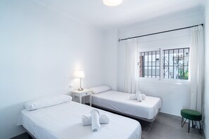 A hotel room with two single beds, a small table with a lamp, a window with bars, and a green stool. - Apartment in Nerja Near Carabeillo Beach (Nerja)