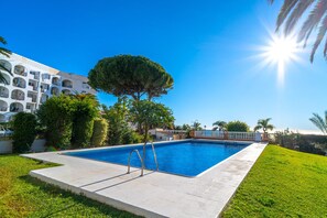 A swimming pool with a clear blue sky, surrounded by greenery and a white building with balconies. - Apartment in Nerja Near Carabeillo Beach (Nerja)