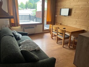 A living room with a sofa, a wooden dining table with chairs, a television mounted on the wall, and a window with curtains. - Comfortable central studio - 4 guests (Pralognan-la-Vanoise)