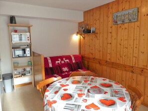 A cozy room with a wooden wall, a bed with a red and white patterned blanket, a wooden shelf with kitchen appliances, and a table with a tablecloth featuring food illustrations. - Well-located studio cabin, cats OK - 4 guests (Pralognan-la-Vanoise)