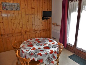 Habitació amb paret de fusta encastrada, taula rodona amb mantell estampat de diari, televisió penjada a la paret i finestra amb cortines.