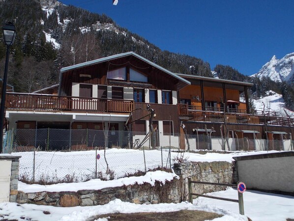 A multi-story wooden building with balconies, a stone retaining wall, and a snow-covered landscape with mountains in the background. - Well-located studio cabin, cats OK - 4 guests (Pralognan-la-Vanoise)