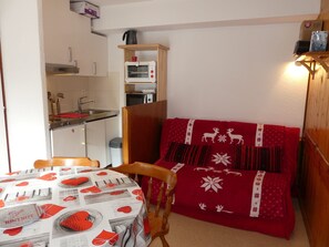 A small kitchenette with a microwave, stove, and sink. There is a dining table with a red tablecloth and a chair. A red bed with reindeer patterns is visible. - Well-located studio cabin, cats OK - 4 guests (Pralognan-la-Vanoise)