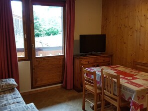 Ein Zimmer mit Holzvertäfelung, einem Fernseher auf einer Holzkommode, einem Esstisch mit Stühlen und einem Fenster mit Vorhängen.