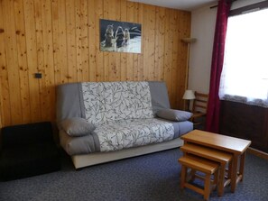 Ein Zimmer mit einer holzverkleideten Wand, einem grauen Sofa mit gemusterten Kissen, einem kleinen Holztisch und einem Fenster mit rotem Vorhang.