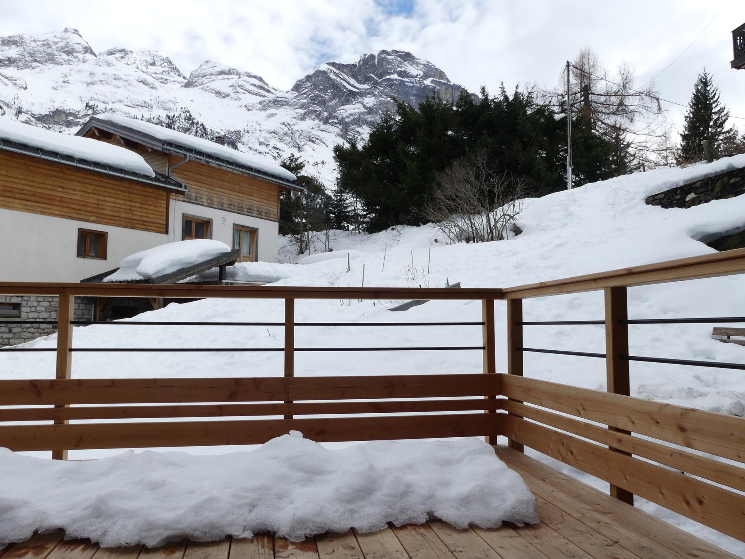 A wooden deck with snow accumulation, overlooking a mountainous landscape with buildings.