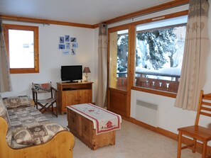 Ein gemütliches Wohnzimmer mit einer Couch, einem Fernseher, einer Holzkommode und einem Fenster mit Blick auf schneebedeckte Bäume.